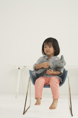 Kleines asiatisches Mädchen mit Stofftier sitzt auf einem Stuhl, lizenzfreies Stockfoto