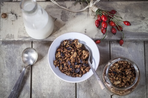 Schale mit Müsli aus gebackenen Haferflocken, Nüssen und Rosinen, Flasche Milch und Hagebutten auf Holzbrett - SBDF000339