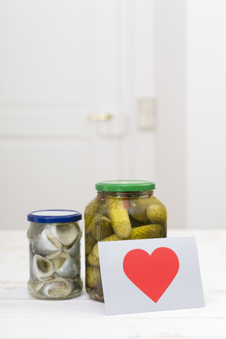 Glass of pickled cucumbers, a glass of rollmops and a postcard with red heart on white wooden table stock photo