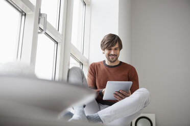 Germany, Munich, Using digital tablet, sitting on window sill - RBF001445