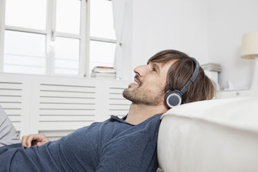 Germany, Munich, Man sitting on sofa, wearing head phones - RBF001482