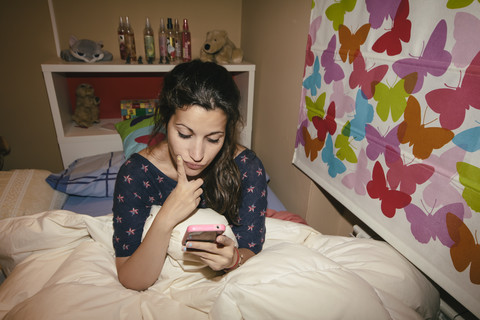 Spanien, Madrid, junge Frau sitzt auf dem Bett und benutzt ein Smartphone, lizenzfreies Stockfoto