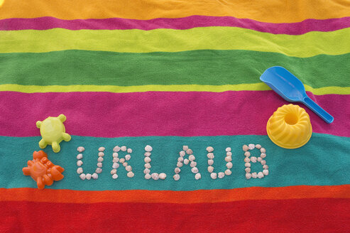 The word 'Urlaub' formed by shells lying between sand toys on a multicolored bath towel - ASF005262