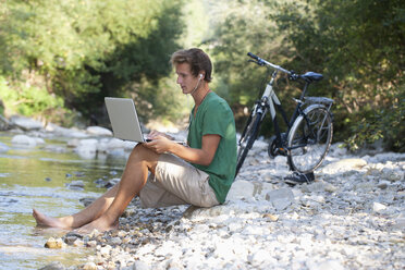 Österreich, Salzkammergut, Mondsee, junger Mann mit Laptop lernt an einem Bach - WWF003189