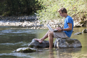 Austria, Salzkammergut, Mondsee, young man with laptop learning at a brook - WWF003190