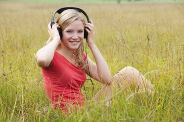Austria, Salzkammergut, Mondsee, young smiling woman with headphones sitting in a meadow - WWF003172