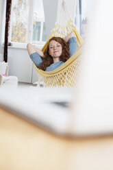 Woman relaxing in a hammock in her apartment, laptop standing in front on a table - RBF001548