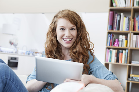 Porträt einer jungen Frau, die sich mit einem Tablet-Computer auf einer Couch in ihrer Wohnung entspannt, lizenzfreies Stockfoto