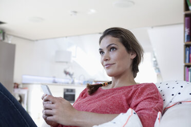 Portrait of woman sitting on a couch in her apartment holding smartphone - RBF001565