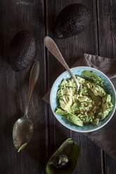 Bowl of crushed avocado, kitchen towel, spoons and whole and hollowed half of avocados on wooden table - SBDF000467