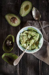 Bowl of crushed avocado, kitchen towel, spoon and sliced and hollowed halfs of two avocados on wooden table - SBDF000468