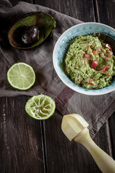 Schüssel mit Guacamole, Küchentuch, Stößel, geschnittene Limette und ausgehöhlte Avocadohälfte auf Holztisch - SBDF000470