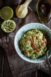 Bowl of guacamole, kitchen towel, pestle, sliced lime and hollowed half of avocado on wooden table - SBDF000471