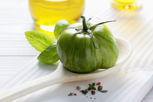 Grüne Tomate auf Holzlöffel, Olivenöl, Basilikum und Pfefferkörner auf Holztisch - CSF020719