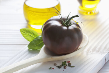 Schwarze Tomate auf Holzlöffel, Olivenöl, Basilikum und Pfefferkörner auf Holztisch - CSF020716