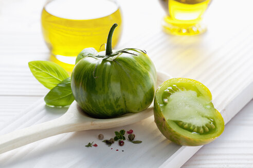 Grüne Tomate auf Holzlöffel, Olivenöl, Basilikum und Pfefferkörner auf Holztisch - CSF020675