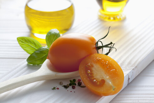 Orangefarbene Tomate auf Holzlöffel, Olivenöl, Basilikum und Pfefferkörner auf Holztisch - CSF020674