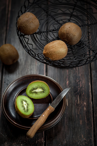 Drahtkorb und Teller mit ganzen und geschnittenen Kiwi-Früchten auf Holztisch, lizenzfreies Stockfoto