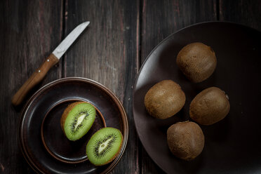 Teller mit ganzen und in Scheiben geschnittenen Kiwi-Früchten und ein Messer auf einem Holztisch - SBDF000490