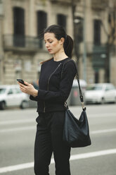 Spain, Catalunya, Barcelona, young black dressed businesswoman with smartphone in front of a street - EBSF000006