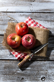 Drei rote Äpfel und ein Messer auf Jute, Stoffserviette und Holztisch - MAEF007768