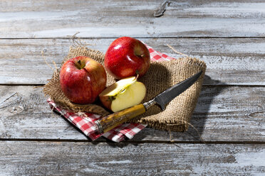 Geschnittene und ganze rote Äpfel und ein Messer auf Jute, Stoffserviette und Holztisch - MAEF007765