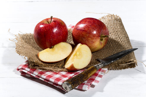 Geschnittene und ganze rote Äpfel und ein Messer auf Jute, Stoffserviette und Holztisch - MAEF007763