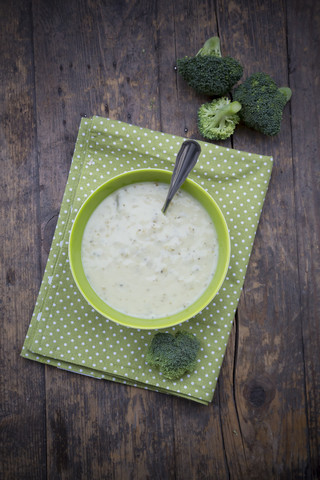 Schüssel mit Brokkolisuppe, Löffel, Tuch und Brokkoliröschen auf Holztisch, lizenzfreies Stockfoto