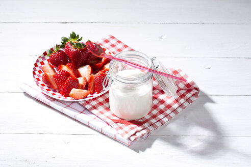 Glas Naturjoghurt mit Plastiklöffel, Teller mit geschnittenen und ganzen Erdbeeren (Fragaria) auf Küchenhandtüchern und weißem Holztisch - MAEF007844