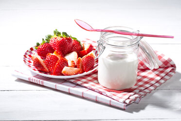 Glas Naturjoghurt mit Plastiklöffel, Teller mit geschnittenen und ganzen Erdbeeren (Fragaria) auf Küchenhandtüchern und weißem Holztisch - MAEF007845