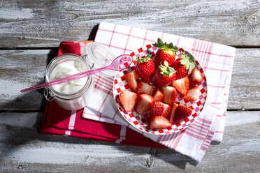 Glas Naturjoghurt mit Plastiklöffel, Teller mit geschnittenen und ganzen Erdbeeren (Fragaria) auf Küchenhandtüchern und Holztisch - MAEF007848
