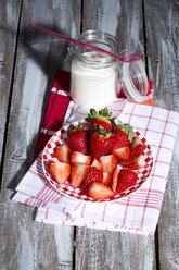 Glas Naturjoghurt mit Plastiklöffel, Teller mit geschnittenen und ganzen Erdbeeren (Fragaria) auf Küchenhandtüchern und Holztisch - MAEF007849