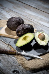 Sliced and whole avocados (Persea americana), baking paper, knife and slate on jute and wooden table - MAEF007852