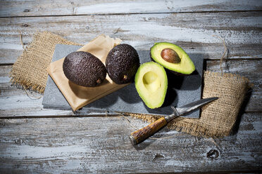 Sliced and whole avocados (Persea americana), baking paper, knife and slate on jute and wooden table - MAEF007819