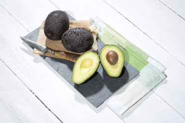 Sliced and whole avocados (Persea americana), baking paper, knife and slate on napkin and white wooden table - MAEF007820