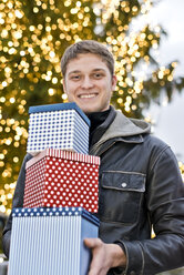 Junger Mann hält drei Geschenkboxen vor einem beleuchteten Weihnachtsbaum - CLPF000068