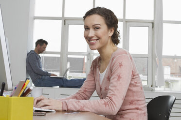 Lächelnde Frau am Schreibtisch mit Mann im Hintergrund - RBYF000384