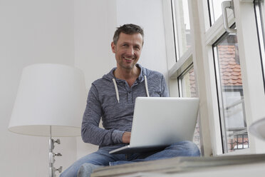 Man sitting on windowsill using laptop - RBYF000386