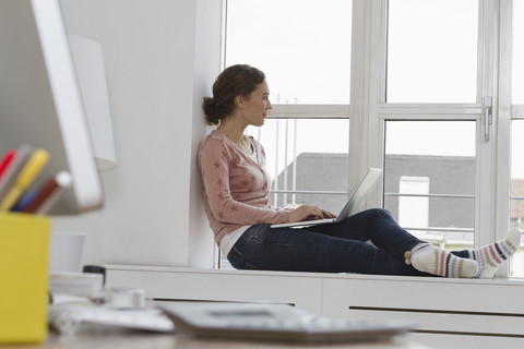 Frau sitzt auf der Fensterbank und benutzt einen Laptop, lizenzfreies Stockfoto