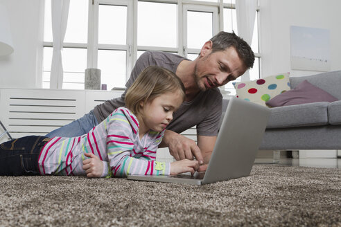 Vater und Tochter benutzen einen Laptop auf dem Teppich im Wohnzimmer - RBYF000485