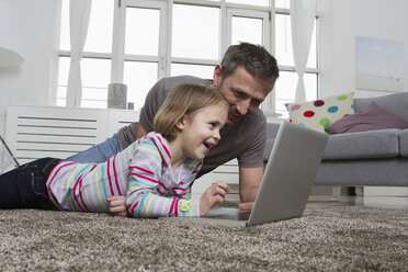 Vater und Tochter benutzen einen Laptop auf dem Teppich im Wohnzimmer - RBYF000486