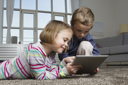 Brother and sister using tablet computer on carpet in living room - RBYF000434