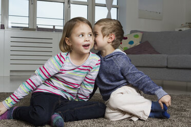 Brother and sister whispering on carpet in living room - RBYF000438