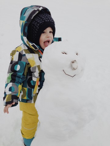 Kleiner Junge im Schnee-Outfit probiert das Eis eines Schneemanns in Reit im Winkl, Bayern, Deutschland, lizenzfreies Stockfoto
