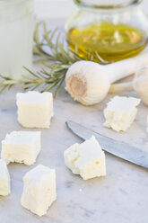 Gewürfelter Schafskäse, ein Glas Olivenöl, Knoblauchzwiebeln, Rosmarin und Messer auf weißem Marmor - LVF000727
