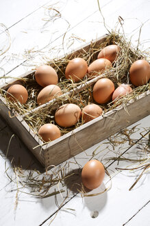 Holzkiste mit braunen Eiern und Heu auf weißem Holztisch - MAEF007948