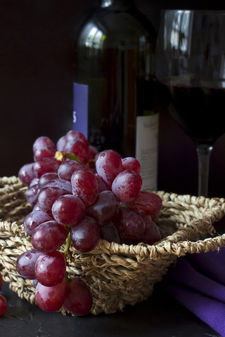 Weidenkorb mit roten Weintrauben (vitis vinifera), Rotweinglas und Flasche im Hintergrund, lizenzfreies Stockfoto