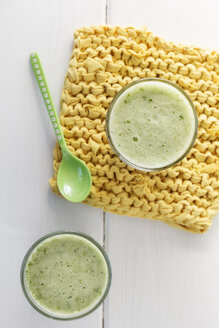 Zwei Gläser Grünkohl-Bananen-Smoothie, gelber Topflappen und Löffel auf weißem Holztisch, Blick von oben - EVGF000400