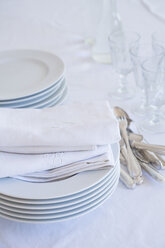 Glasses, stack of plates, silver cutlery and cloth napkins on white table cloth - LVF000778