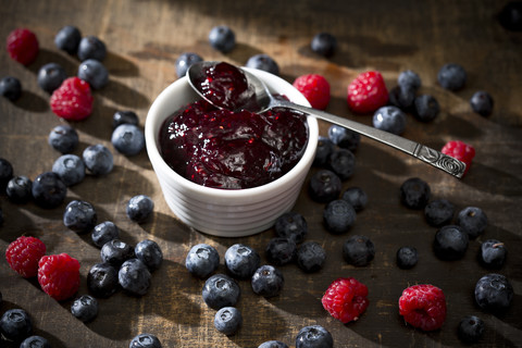 Schale mit Heidelbeer- und Himbeermarmelade, Löffel, Heidelbeeren und Himbeeren auf dunklem Holztisch, lizenzfreies Stockfoto